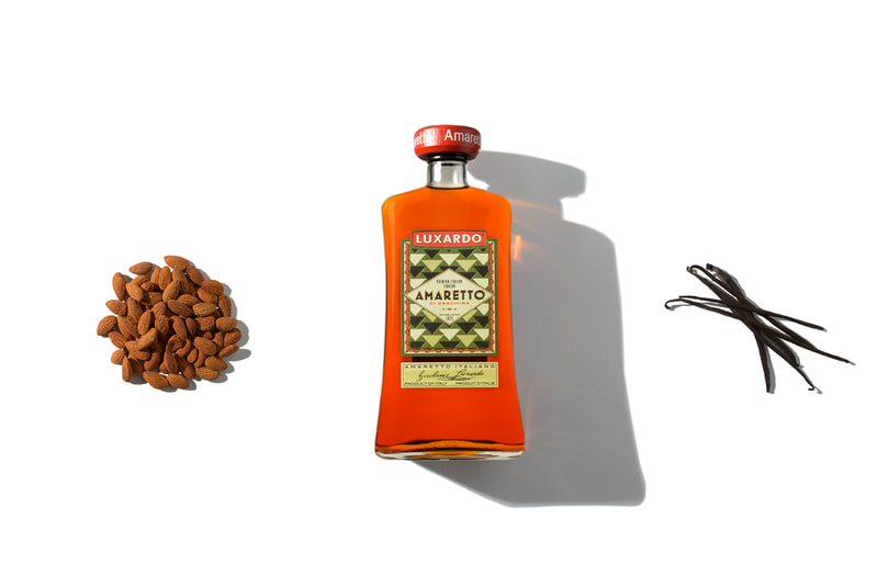 Three objects are brightly lit as they lay on a pure white surface. A mound of peeled almonds on the left, a bundle of whole vanilla beans on the right and a bottle of Luxardo Amaretto liquor in the middle.