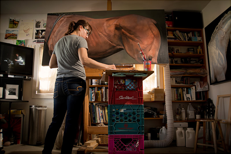 A female artist stands in front of her large painting of a horse.