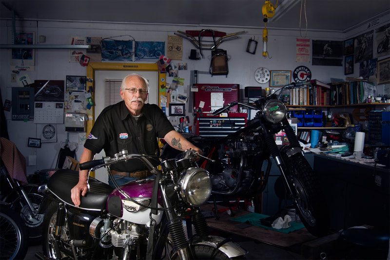 A silver haired gentlemen is shown in his home workshop, he stands sturdy behind his purple motorcycle.