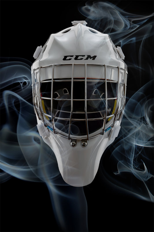 A white hockey goalie helmet is photographed against a black background. The helmet is engulfed in a fine swirl of blue-white smoke. The feeling is ominous and mysterious. Shot by photographer Mike Taylor of Peterborough Ontario.