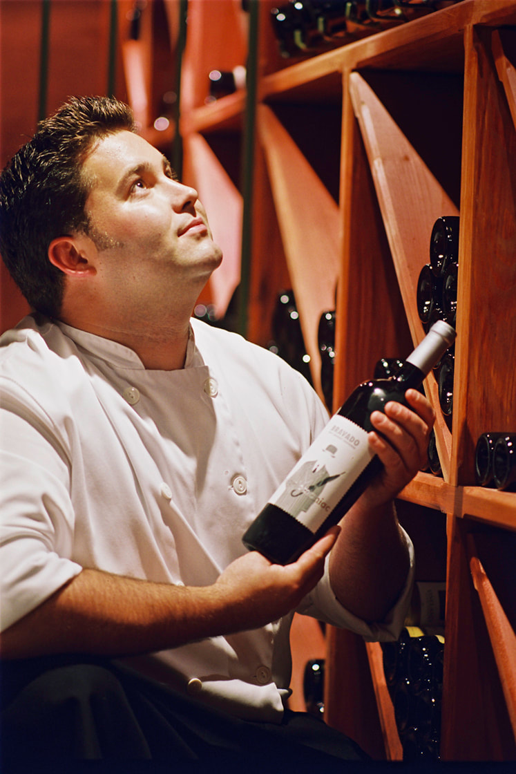 In this wine cellar scene, a chef in white clothing, holds a bottle of red wine. He looks upwards to see what other wines are available.