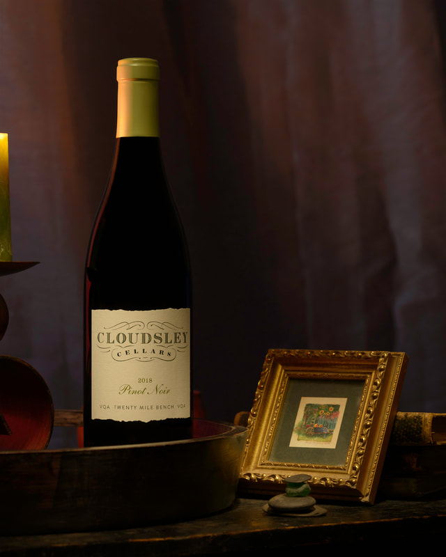 A bottle of red with with a white label sits on a dark table straddled between a small gold picture from and a curvaceous candle holder.