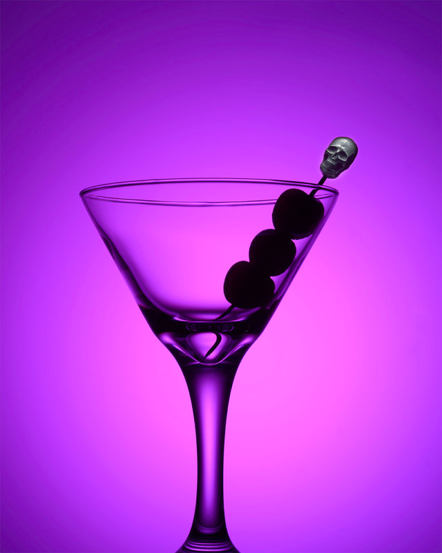 A bright purple scene occupied by a single martini glass. Inside is a garnish pick topped with a metallic skull and 3 cherries skewered upon.
