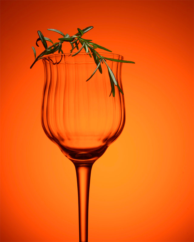 A fluted wine glass with a rosemary garnish, sits in front of a orange background