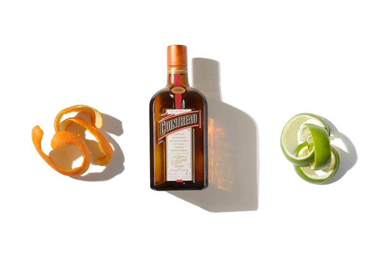 Three objects are brightly lit as they lay on a pure white surface. A curled orange peel on the left, a curled lime peel on the right and a bottle of Cointreau liquor in the middle.