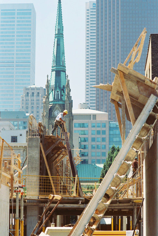 A construction worker is shown from a distance, looking down from a great height. The cityscape of towers looms in the background