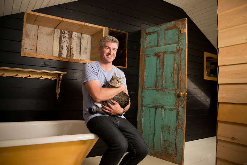 A handsome man sits on the edge of an antique bathtub. He holds a cat.