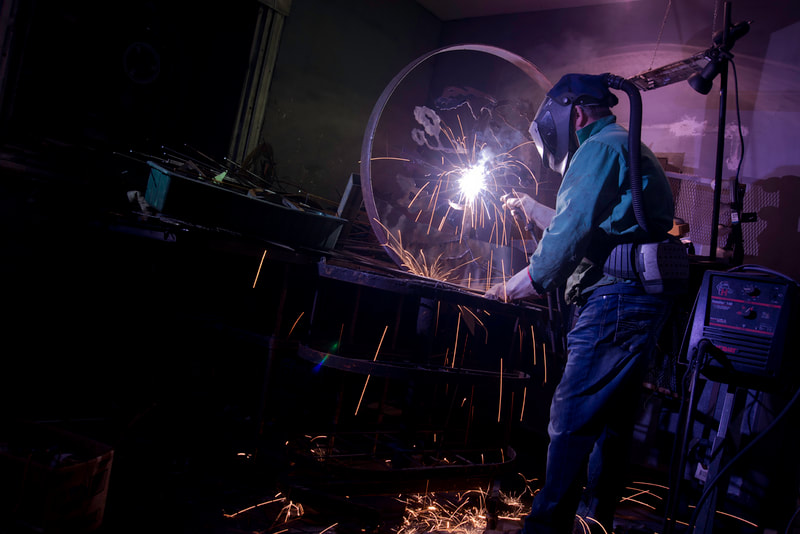 An artisan working in metal is shown using a welding instrument on a large circular form.