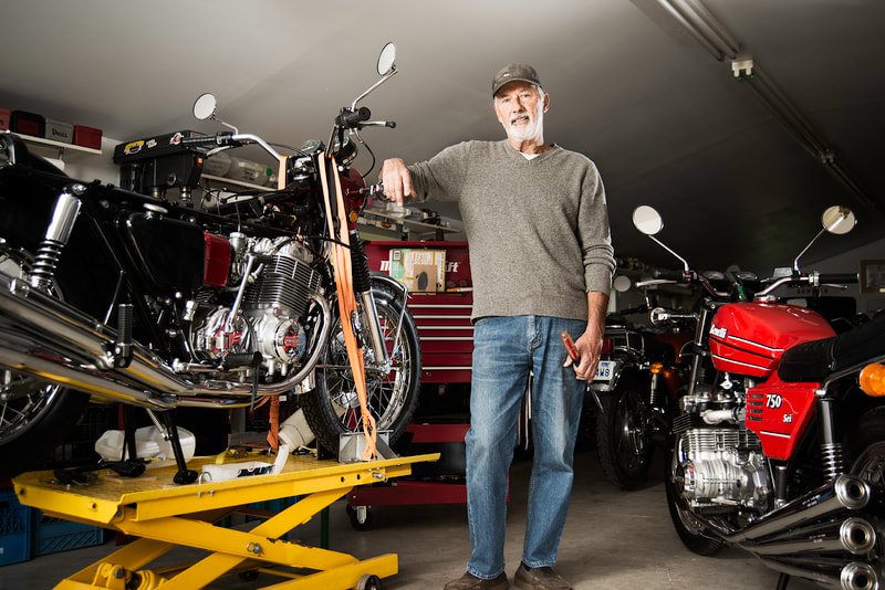 A tall bearded man stands in a workshop. He holds a tool in his left hand, while he leans on a motorcycle with his right arm.