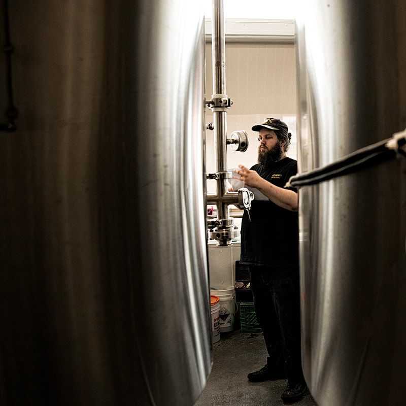 A bearded man is framed by two ominous steel tanks as he examines a measuring device in a brewery.