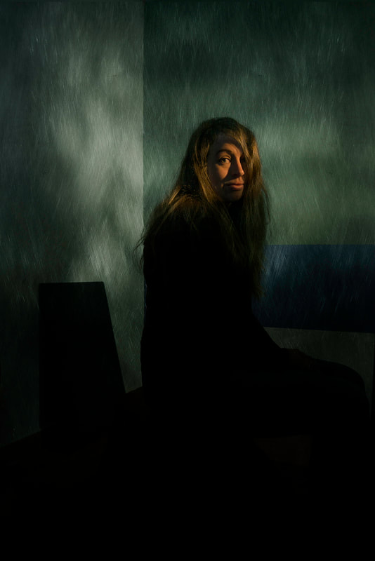 A dramatic portrait of a woman with long hair. She sits in a dark room with only a gentle light illuminating her face.