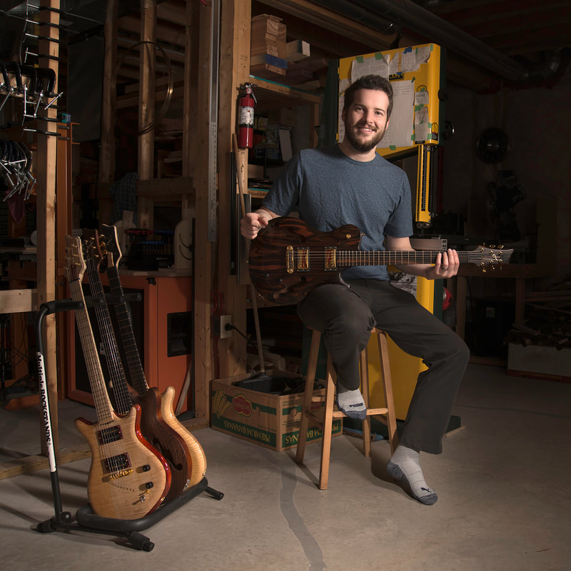 A young bearded man sits on a stool in a workshop. He holds a guitar that he made while smiling at the camera.