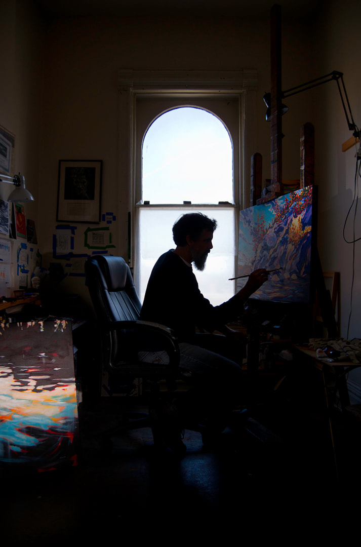 A bearded man is silhouetted against a tall window. He is painting a colourful canvas.