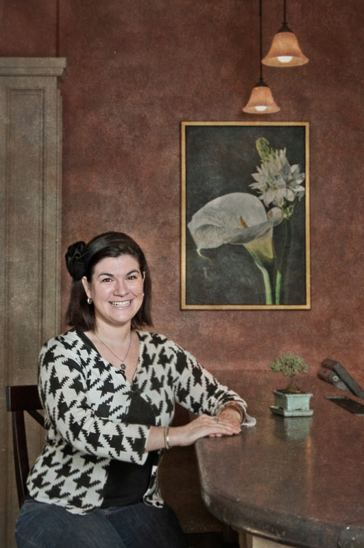 A smiling woman sits at a  restaurant bar. Behind her is a framed painting of a Cala lily. Image shot by photographer Mike Taylor in Peterborough Ontario.