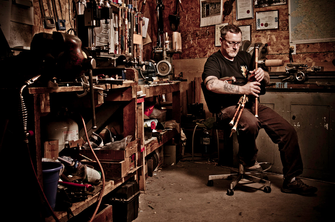 A man sits on a stool in a crowded workshop as he plays a set of Irish bagpipes.