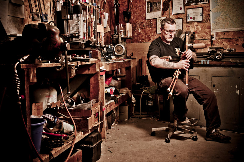 A bagpipe player sits in a workshop, playing the pipes.