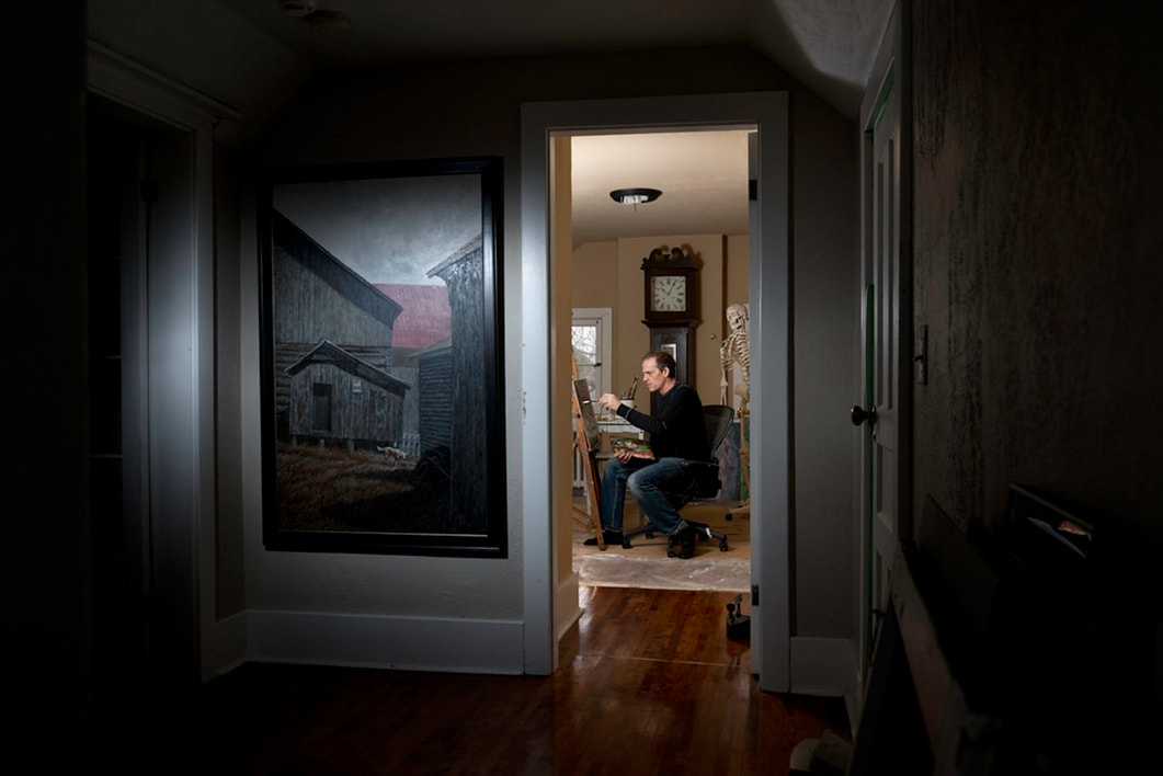 A man in a black shirt and blue jeans sits in front of a canvas and paints a picture.