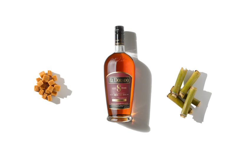 Three objects are brightly lit as they lay on a pure white surface. A mound of caramel candies on the left, a pile of sugar cane on the right and a bottle of 8 year old dark rum in the middle.