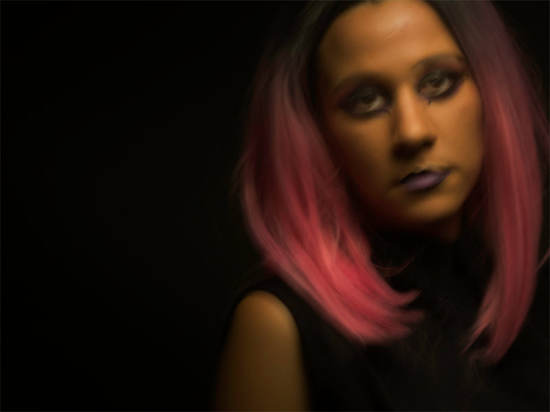 A young woman with pink hair sits in a blackened room. She is not in sharp focus.