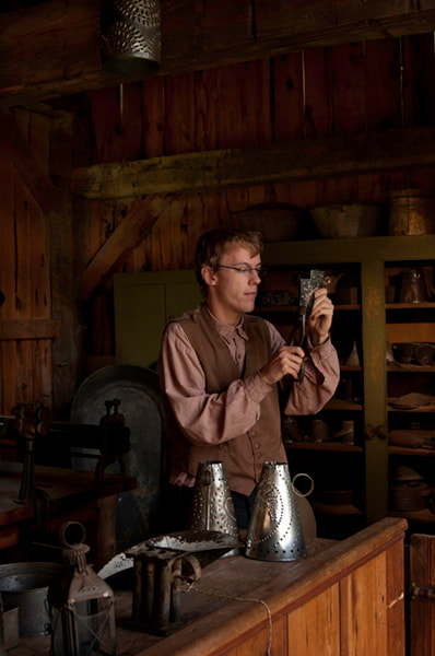 In a recreation of a century old workshop a young bespectacled man holds a shiny metal form in front of his face to examine his work.