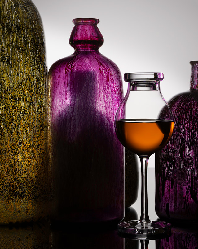 A colourful grouping of handblown glass bottles surround a single whisky glass. Shot in the studio by Peterborough photographer Mike Taylor.