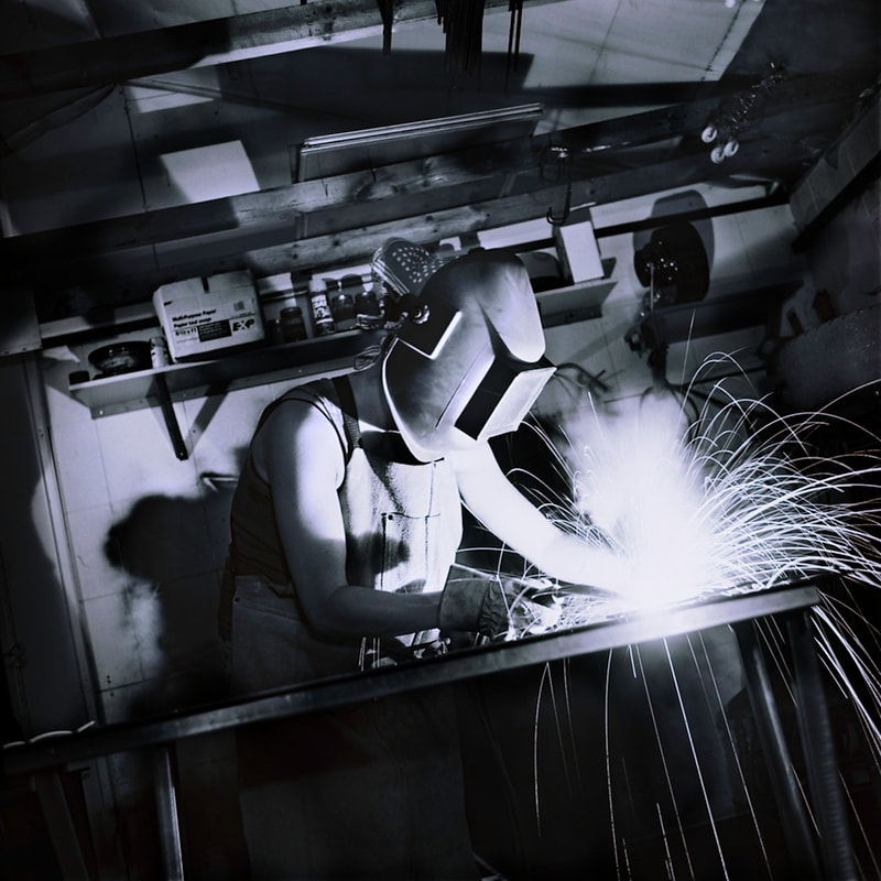 A black and white scene shows a person wearing a welding helmet. Sparks fly aggressively from their work.