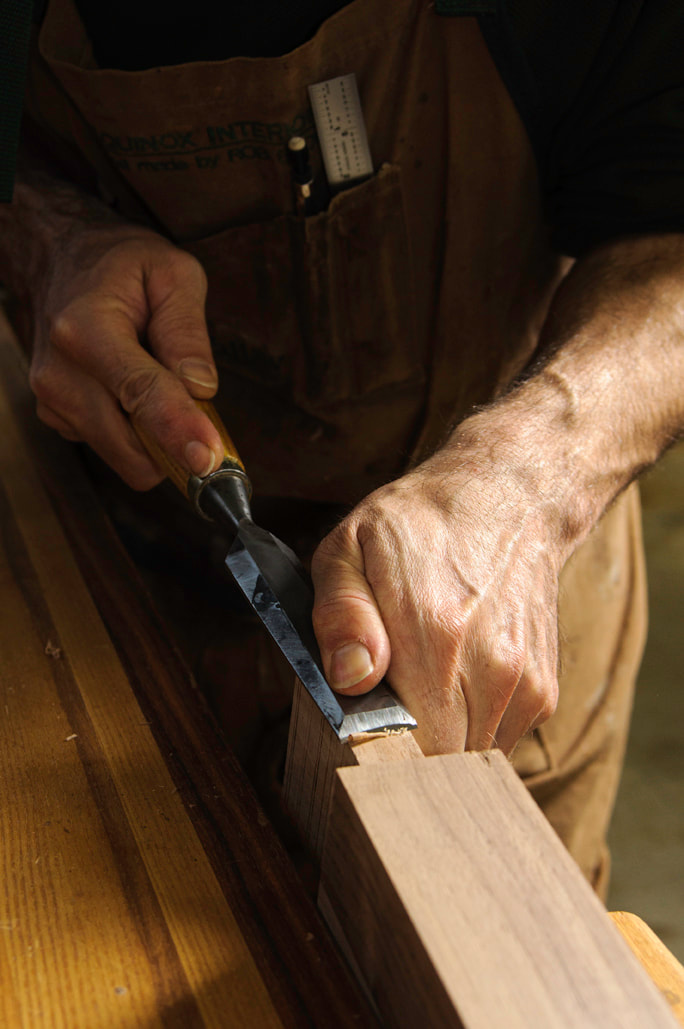 The hands of a woodworker are shown using a fine chisel on some wood.